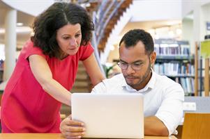 Instructor helping trainee with project in library. Man and woman in casual sitting and standing at desk, using laptop, pointing at monitor. Tutor concept